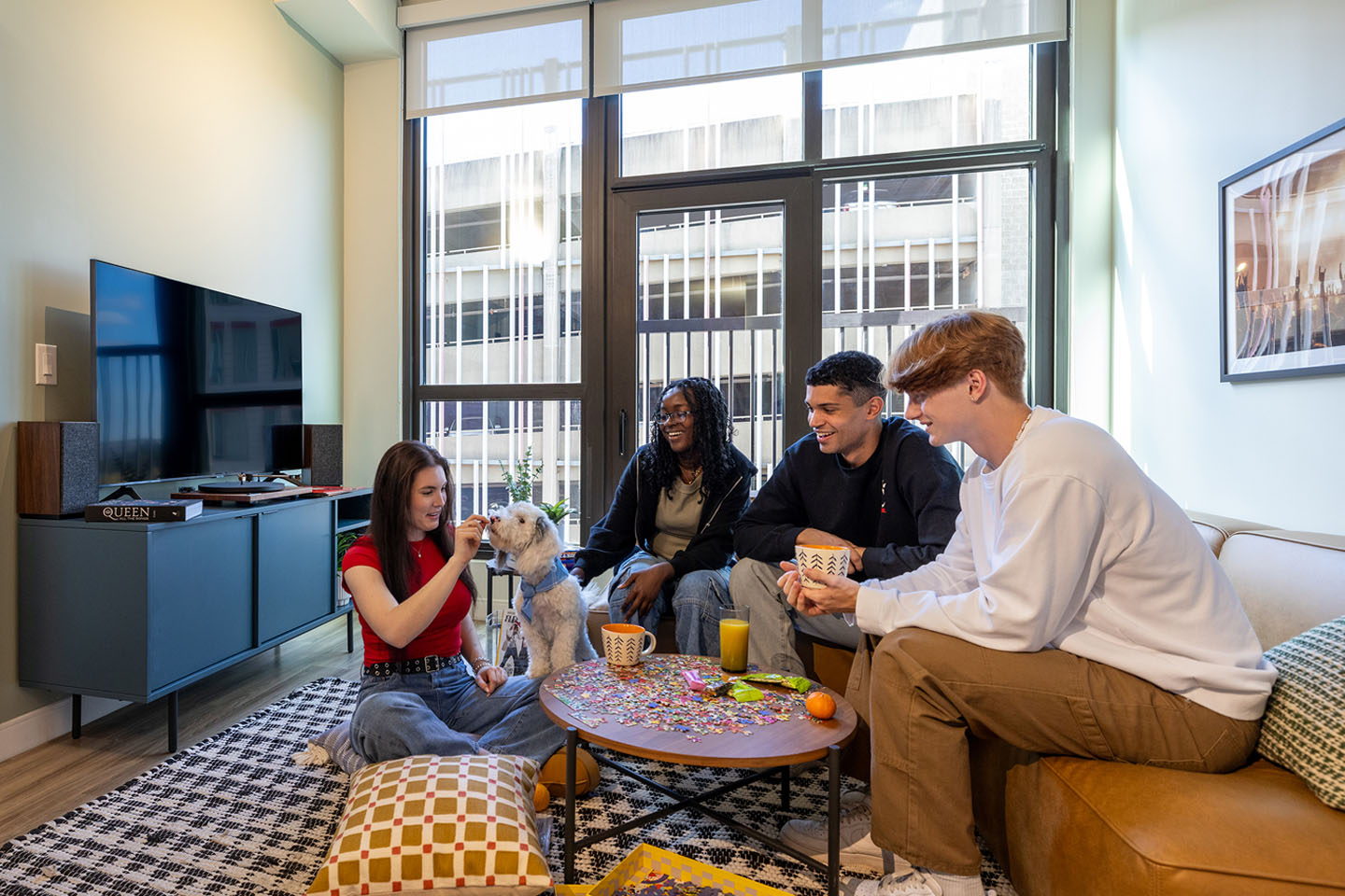 Four young adults hanging out in an apartment with a white long-haired dog.